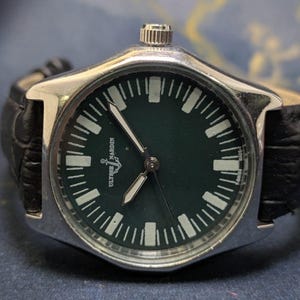 May include: A silver-toned wristwatch with a dark green face and a black leather band. The watch face features white hour markers and hands, with the brand name "Ulysse Nardin" visible. The watch has a classic, sporty design.