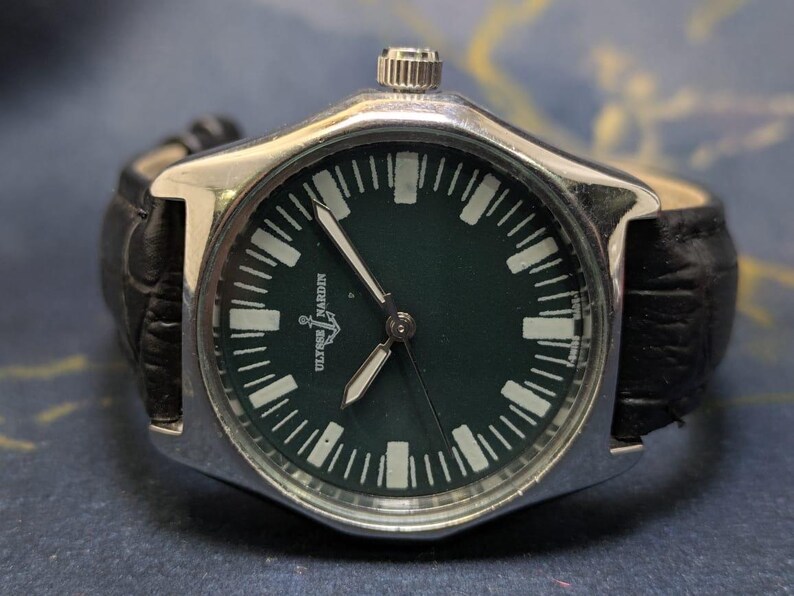 May include: A silver-toned wristwatch with a dark green face and a black leather band. The watch face features white hour markers and hands, with the brand name "Ulysse Nardin" visible. The watch has a classic, sporty design.
