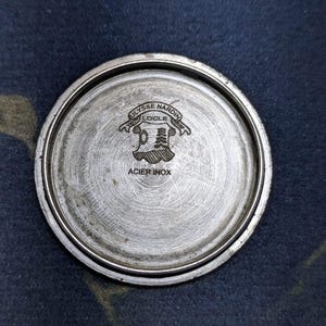 May include: A circular, brushed metal watch back with the engraved text "ULYSSE NARDIN LOCLE" above a crest design. The words "ACIER INOX" are also engraved on the watch back.