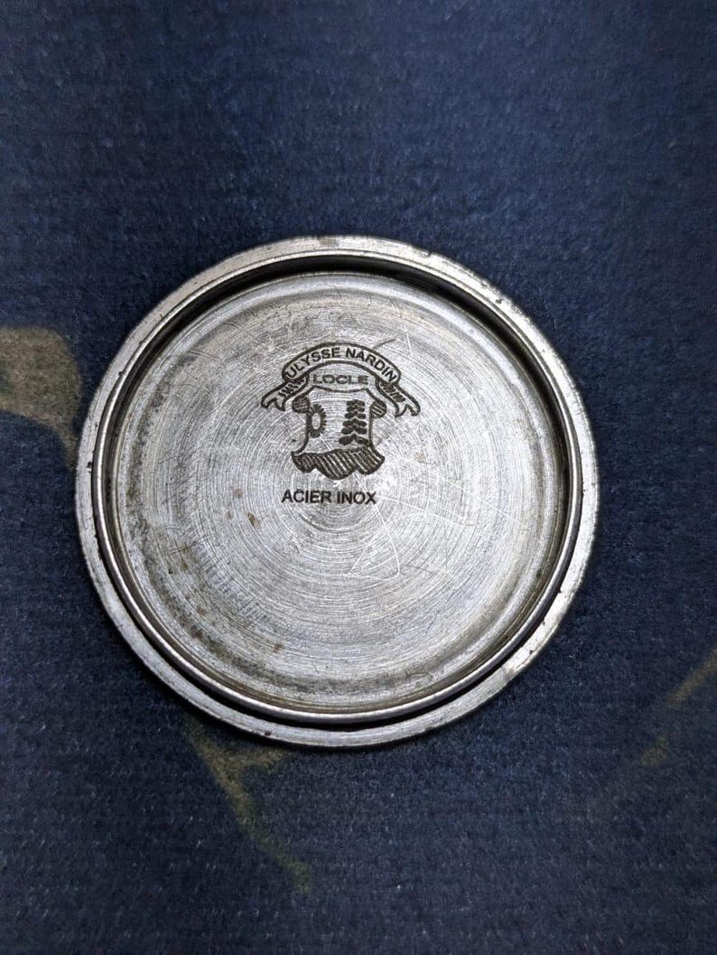 May include: A circular, brushed metal watch back with the engraved text "ULYSSE NARDIN LOCLE" above a crest design. The words "ACIER INOX" are also engraved on the watch back.