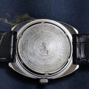 May include: A silver-toned wristwatch with a black leather band. The watch face is not visible. The back of the watch has a circular, textured design. The watch is set against a dark blue background.