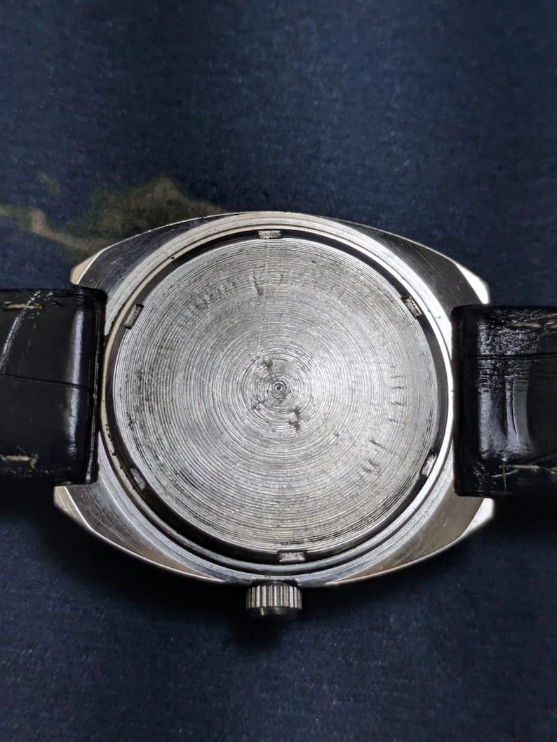 May include: A silver-toned wristwatch with a black leather band. The watch face is not visible. The back of the watch has a circular, textured design. The watch is set against a dark blue background.