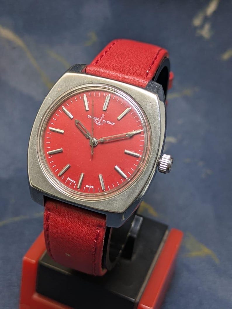 May include: A vintage Ulysse Nardin watch with a square silver case and a vibrant red dial. The watch features silver hands and markers, and a red leather strap. The watch is marked "Swiss Made".