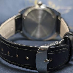 May include: Close-up of a wristwatch with a black leather band and a silver-colored buckle. The watch's back is visible, showing a circular, textured design. The band is attached to the watch with a light-colored material.