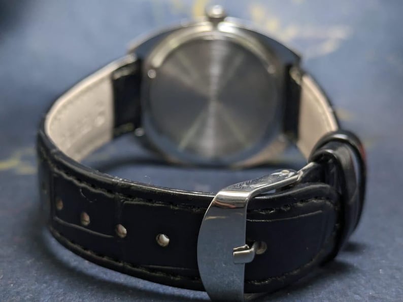 May include: Close-up of a wristwatch with a black leather band and a silver-colored buckle. The watch's back is visible, showing a circular, textured design. The band is attached to the watch with a light-colored material.