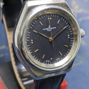 May include: A vintage Ulysse Nardin watch with a black face and gold-tone hands and markers. The watch has a silver-tone case and a black leather band. The watch face features the brand name and the word "SWISS MADE".