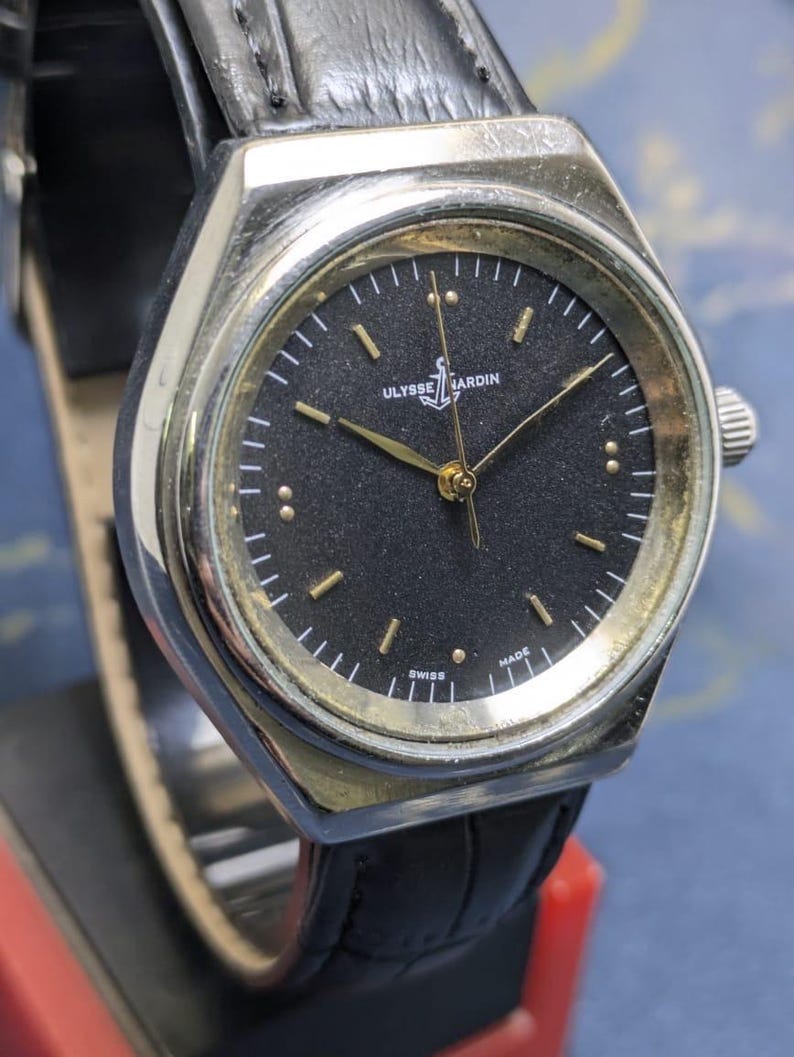 May include: A vintage Ulysse Nardin watch with a black face and gold-tone hands and markers. The watch has a silver-tone case and a black leather band. The watch face features the brand name and the word "SWISS MADE".