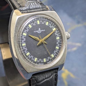 May include: A vintage Ulysse Nardin wristwatch with a black dial and gold hands. The watch has a silver-toned square case and a black leather band. The dial features the Ulysse Nardin logo and the words "SWISS MADE".