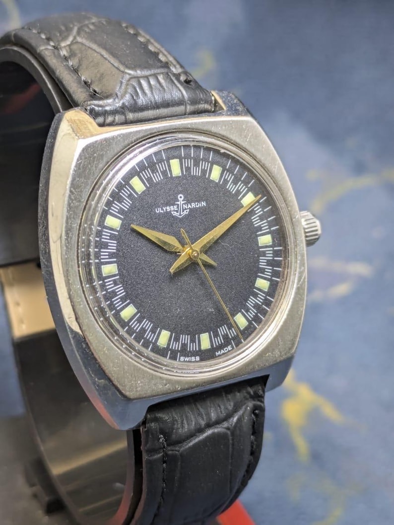 May include: A vintage Ulysse Nardin wristwatch with a black dial and gold hands. The watch has a silver-toned square case and a black leather band. The dial features the Ulysse Nardin logo and the words "SWISS MADE".