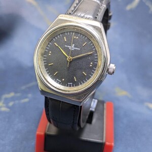 May include: A silver-toned Ulysse Nardin watch with a black face and gold hands. The watch has a black leather band and a silver crown. The watch is displayed on a red and black stand.