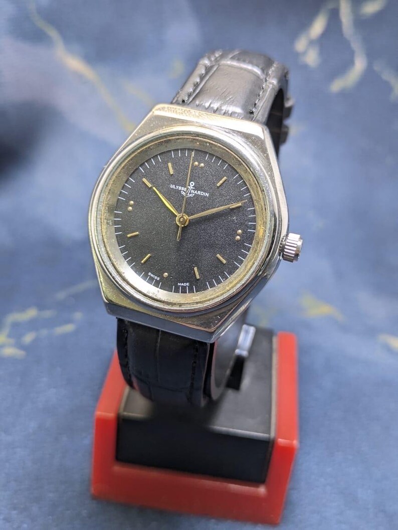 May include: A silver-toned Ulysse Nardin watch with a black face and gold hands. The watch has a black leather band and a silver crown. The watch is displayed on a red and black stand.