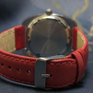 May include: A close-up shot of a wristwatch featuring a red leather strap. The watch has a silver-toned case and a stainless steel buckle. The rear of the watch is visible, displaying a circular pattern. The strap is stitched with a contrasting thread.