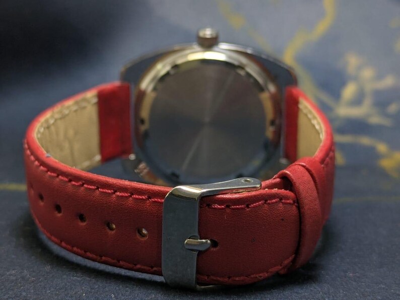 May include: A close-up shot of a wristwatch featuring a red leather strap. The watch has a silver-toned case and a stainless steel buckle. The rear of the watch is visible, displaying a circular pattern. The strap is stitched with a contrasting thread.