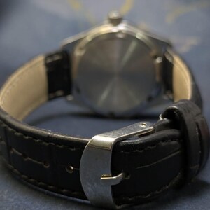May include: Close-up of a wristwatch with a black leather band and a silver buckle. The watch face is visible in the background, with a silver casing. The band is textured and stitched.