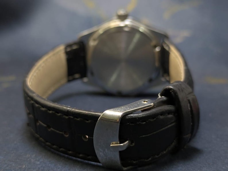 May include: Close-up of a wristwatch with a black leather band and a silver buckle. The watch face is visible in the background, with a silver casing. The band is textured and stitched.