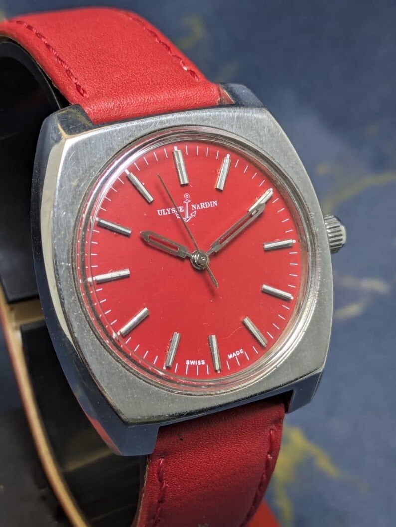 May include: A vintage wristwatch with a square silver case and a vibrant red dial. The watch features silver hands and markers, a red leather strap, and the brand name "Ulysse Nardin". The watch is marked "Swiss Made".