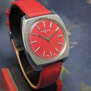 May include: A vintage Ulysse Nardin watch featuring a striking red face and a coordinating red leather strap. The watch has a silver-toned square case and silver hands. The watch is displayed on a black and red stand.