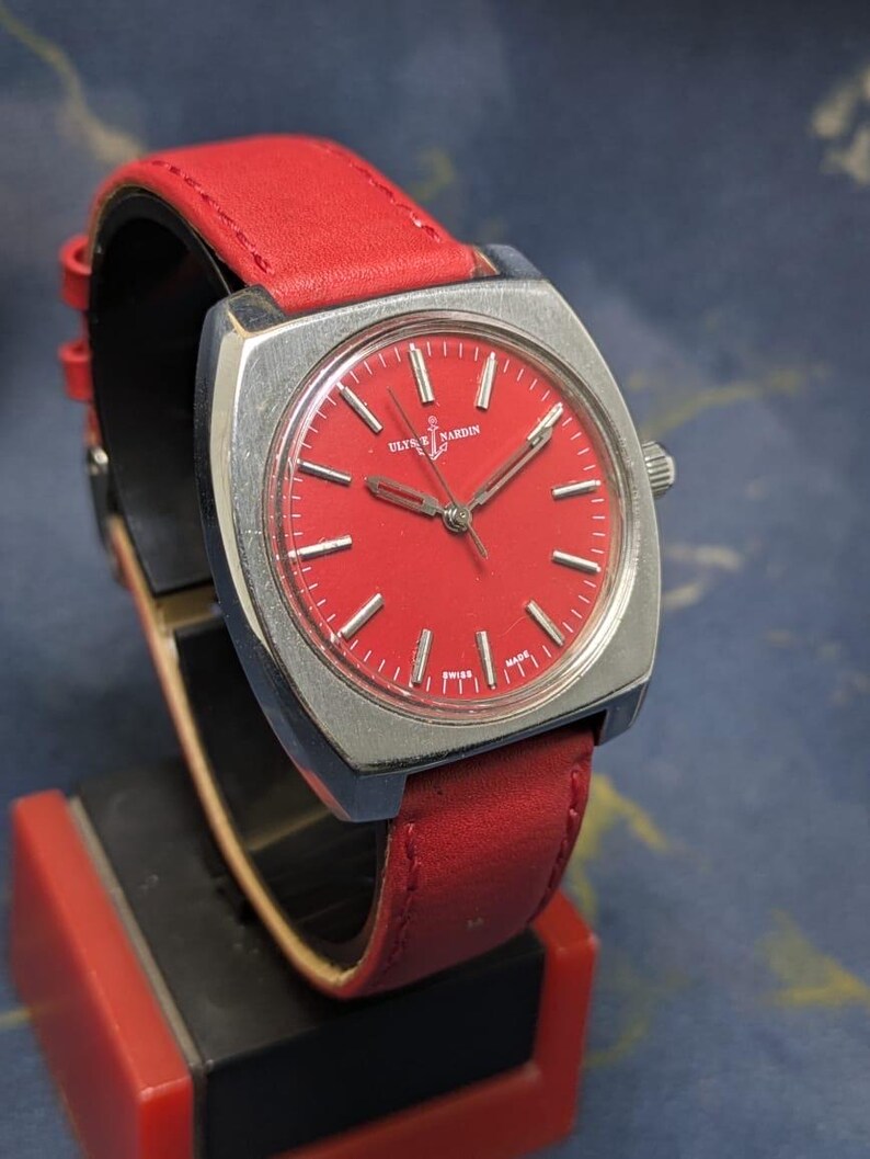 May include: A vintage Ulysse Nardin watch featuring a striking red face and a coordinating red leather strap. The watch has a silver-toned square case and silver hands. The watch is displayed on a black and red stand.