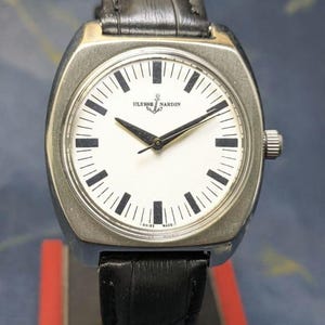 May include: A silver-toned Ulysse Nardin wristwatch with a white face, black hands, and black rectangular hour markers. The watch has a black leather band and a silver-toned case. The watch face displays the Ulysse Nardin logo and the words "SWISS MADE."
