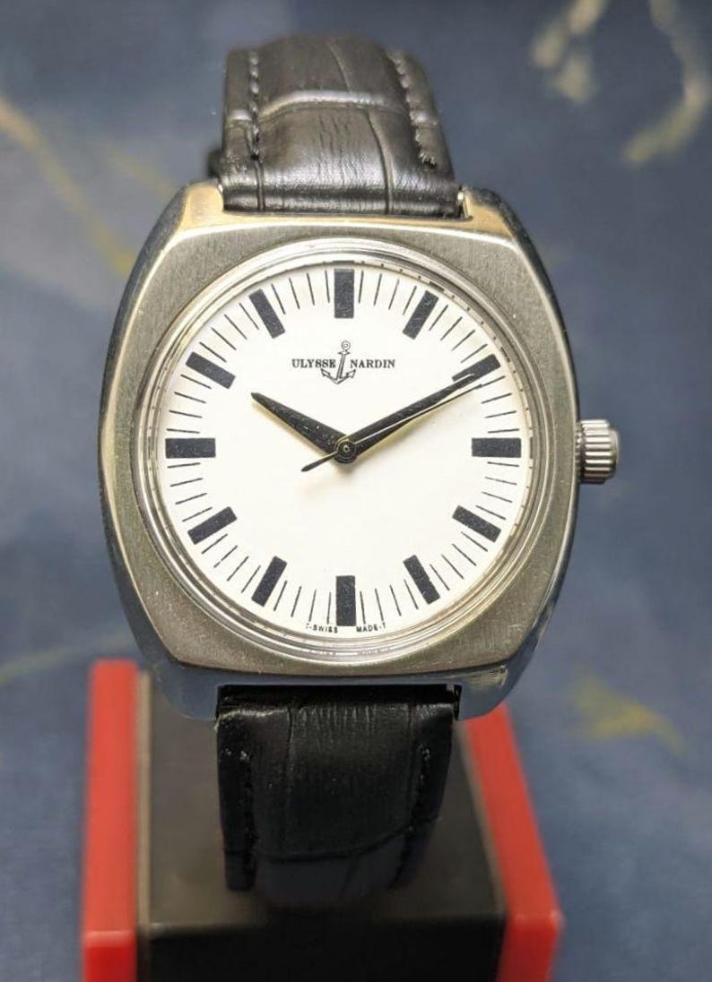 May include: A silver-toned Ulysse Nardin wristwatch with a white face, black hands, and black rectangular hour markers. The watch has a black leather band and a silver-toned case. The watch face displays the Ulysse Nardin logo and the words "SWISS MADE."