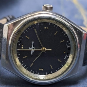 May include: A vintage Ulysse Nardin wristwatch with a black dial and gold-tone accents. The watch has a silver-tone case and a black leather band. The watch face features gold hands and markers. The watch is marked "Swiss Made".