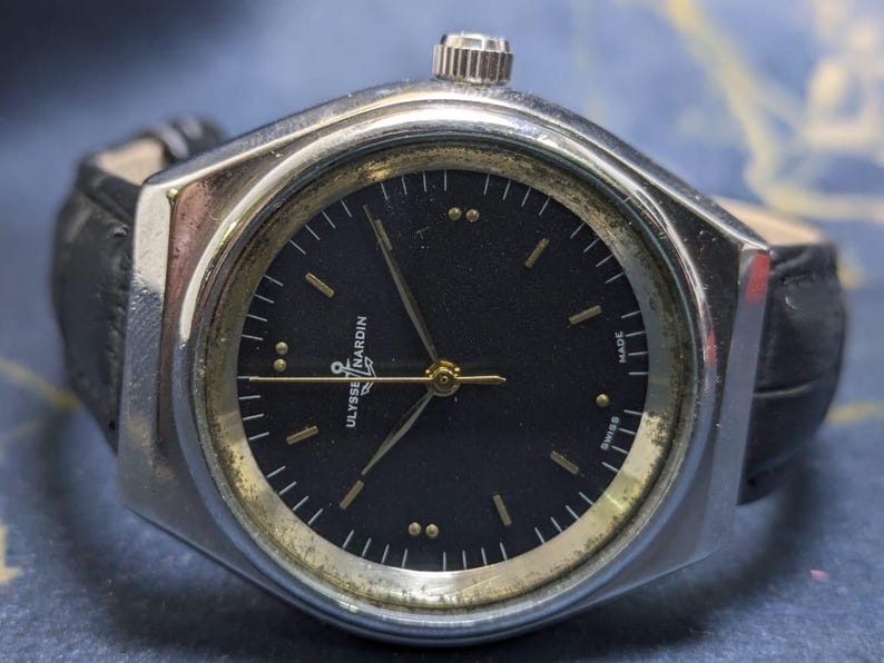 May include: A vintage Ulysse Nardin wristwatch with a black dial and gold-tone accents. The watch has a silver-tone case and a black leather band. The watch face features gold hands and markers. The watch is marked "Swiss Made".