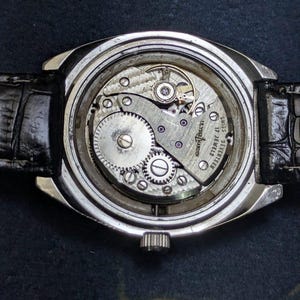 May include: Close-up of a vintage wristwatch with a silver case and a black leather band. The watch's inner workings are visible, showcasing intricate gears and the inscription "SWISS SEVENTEEN 17 JEWELS".