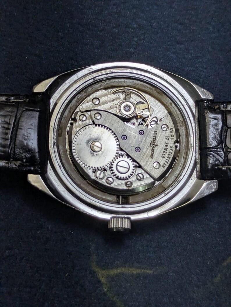 May include: Close-up of a vintage wristwatch with a silver case and a black leather band. The watch's inner workings are visible, showcasing intricate gears and the inscription "SWISS SEVENTEEN 17 JEWELS".