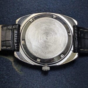 May include: Close-up of the back of a silver-toned wristwatch with a black leather band. The watch face is circular with a textured, brushed metal finish. The band has a crocodile skin pattern.