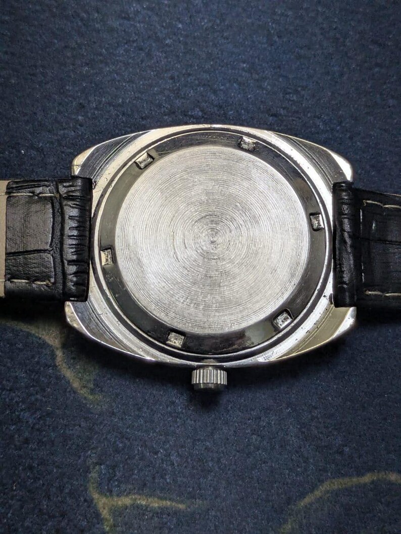 May include: Close-up of the back of a silver-toned wristwatch with a black leather band. The watch face is circular with a textured, brushed metal finish. The band has a crocodile skin pattern.