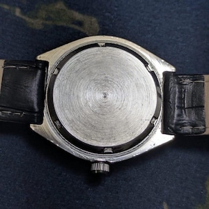 May include: Close-up of the back of a silver-toned wristwatch with a black leather band. The watch face is circular with a textured, metallic finish. The band is attached to the watch with black leather straps.