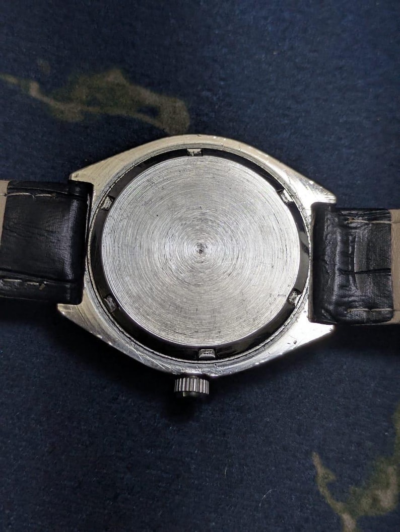 May include: Close-up of the back of a silver-toned wristwatch with a black leather band. The watch face is circular with a textured, metallic finish. The band is attached to the watch with black leather straps.