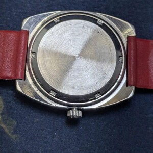 May include: A close-up of the reverse side of a vintage wristwatch. The watch features a silver-toned, rectangular case with a textured, circular back. A red leather strap is attached. The watch crown is visible.