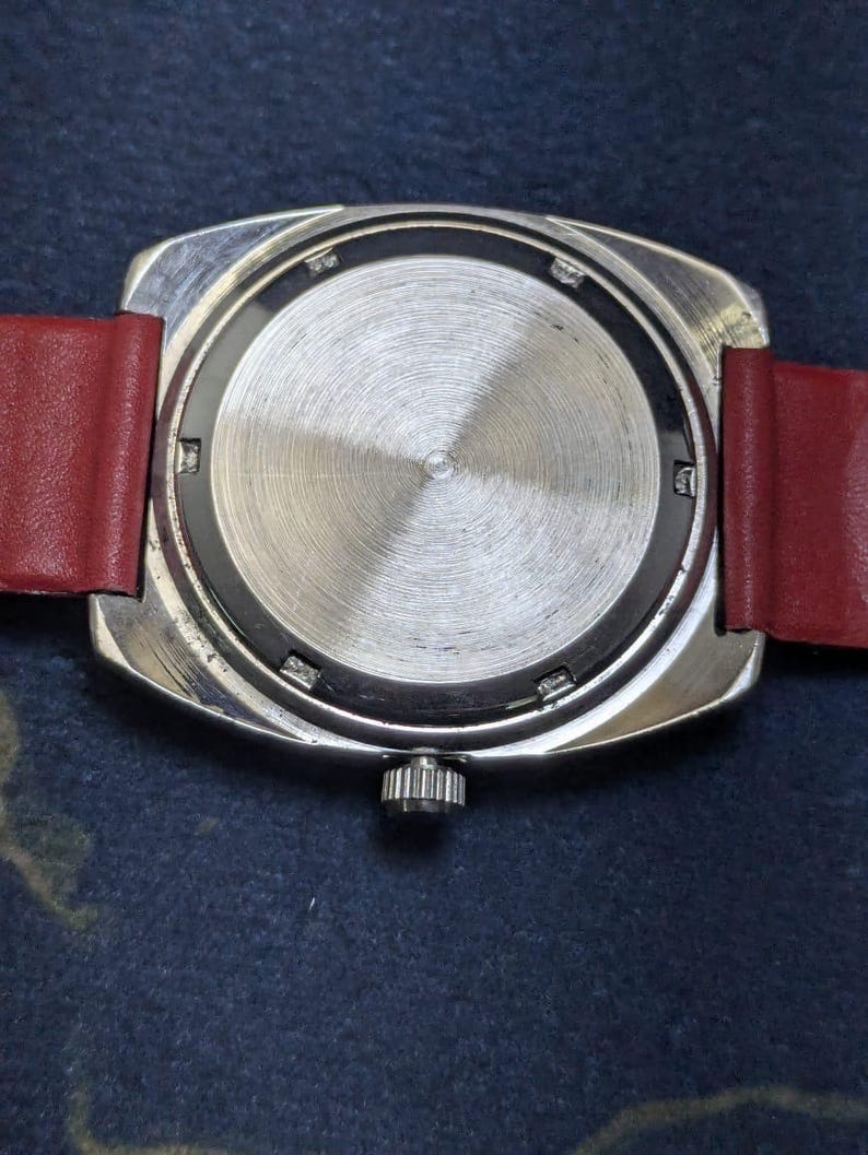 May include: A close-up of the reverse side of a vintage wristwatch. The watch features a silver-toned, rectangular case with a textured, circular back. A red leather strap is attached. The watch crown is visible.
