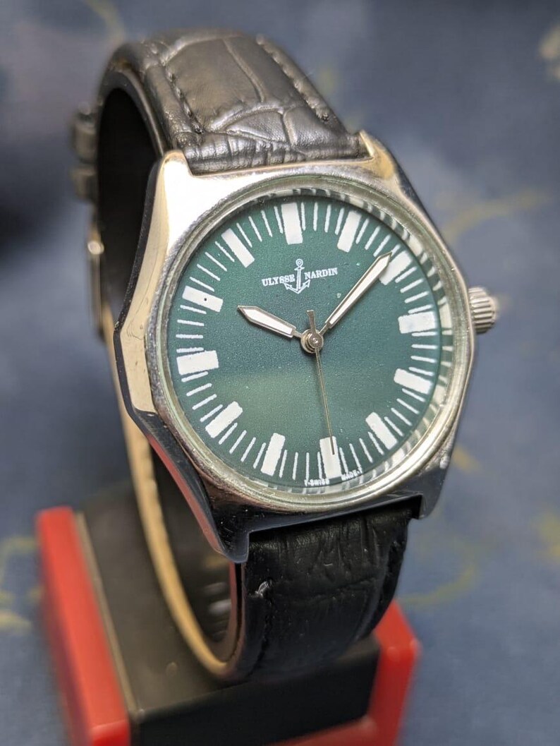 May include: A silver-toned Ulysse Nardin wristwatch with a dark green face and white markers. The watch has a black leather band. The watch face features the brand name and an anchor symbol. The watch is displayed on a black and red stand.