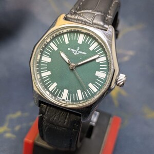 May include: A silver-toned wristwatch with a dark green face and a black leather band. The watch face features white markers and hands, with the brand name "ULYSSE NARDIN" and an anchor logo. The watch is displayed on a red and black stand.