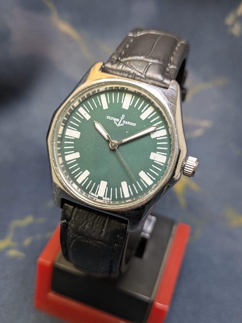 May include: A silver-toned wristwatch with a dark green face and a black leather band. The watch face features white markers and hands, with the brand name "ULYSSE NARDIN" and an anchor logo. The watch is displayed on a red and black stand.