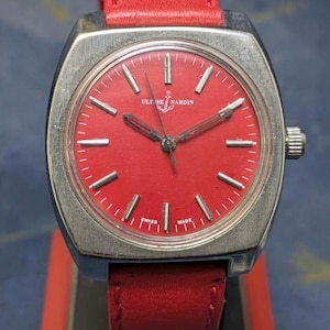 May include: A vintage Ulysse Nardin wristwatch with a bright red face and a red leather strap. The watch has a silver-coloured case and hands, with the brand name and anchor logo on the dial. The watch is marked "Swiss Made".