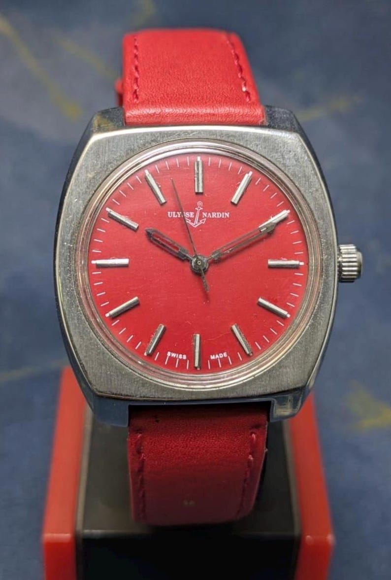 May include: A vintage Ulysse Nardin wristwatch with a bright red face and a red leather strap. The watch has a silver-coloured case and hands, with the brand name and anchor logo on the dial. The watch is marked "Swiss Made".