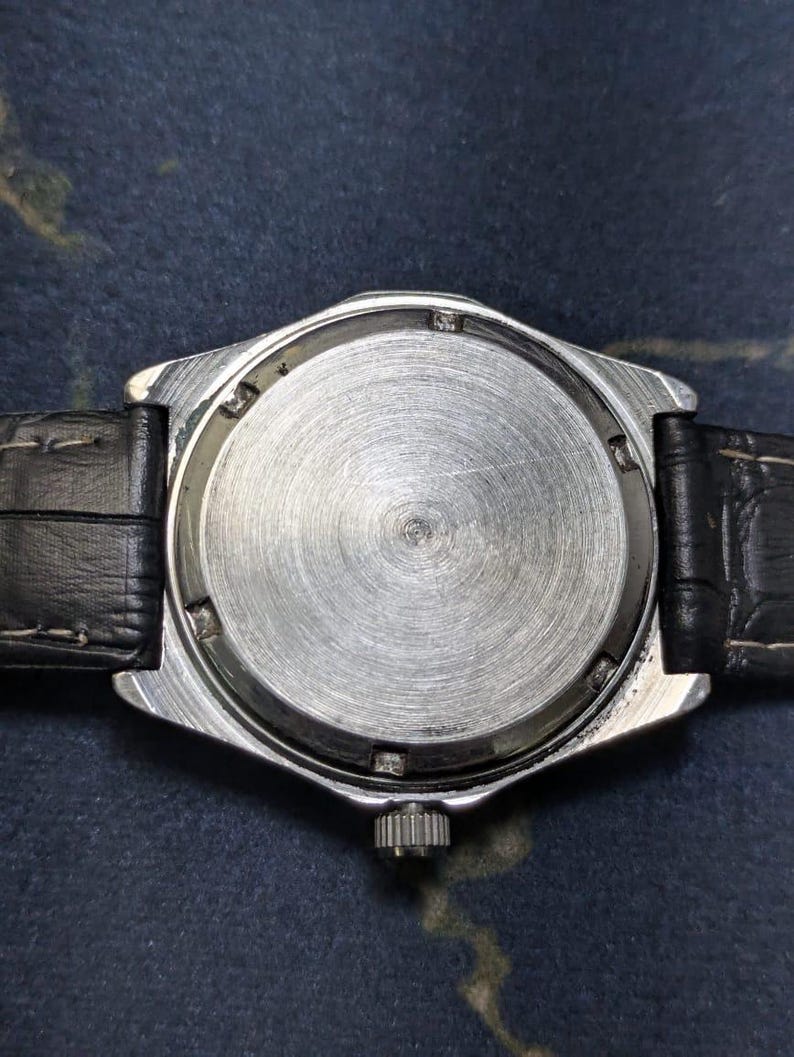 May include: Close-up of the back of a silver-toned wristwatch with a black leather band. The watch face is a brushed metal with a circular design. The band is black and textured.
