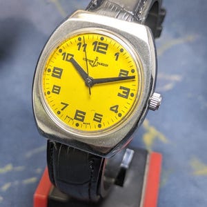 May include: A vintage Ulysse Nardin watch with a bright yellow face and black numerals. The watch has a silver-toned case and a black leather band. The watch face displays the brand name and the words "Swiss Made".