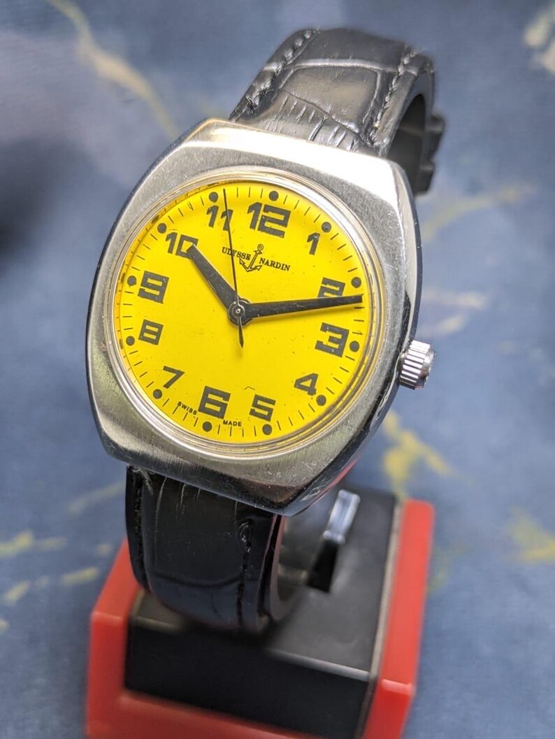 May include: A vintage Ulysse Nardin watch with a bright yellow face and black numerals. The watch has a silver-toned case and a black leather band. The watch face displays the brand name and the words "Swiss Made".