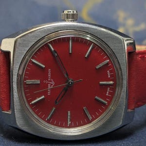 May include: A vintage Ulysse Nardin wristwatch with a bright red dial and a silver-coloured case. The watch has a red leather strap and a silver crown. The dial features silver markers and hands. The watch is marked "SWISS MADE".
