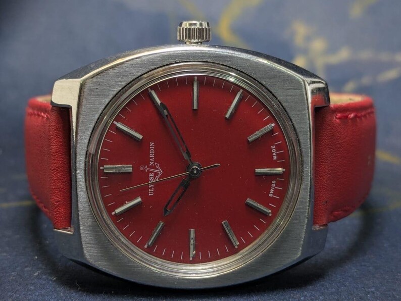 May include: A vintage Ulysse Nardin wristwatch with a bright red dial and a silver-coloured case. The watch has a red leather strap and a silver crown. The dial features silver markers and hands. The watch is marked "SWISS MADE".
