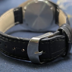 May include: Close-up of a black leather watch strap with a silver buckle. The watch strap is textured and stitched, with a light-colored interior. The watch face is visible in the background.