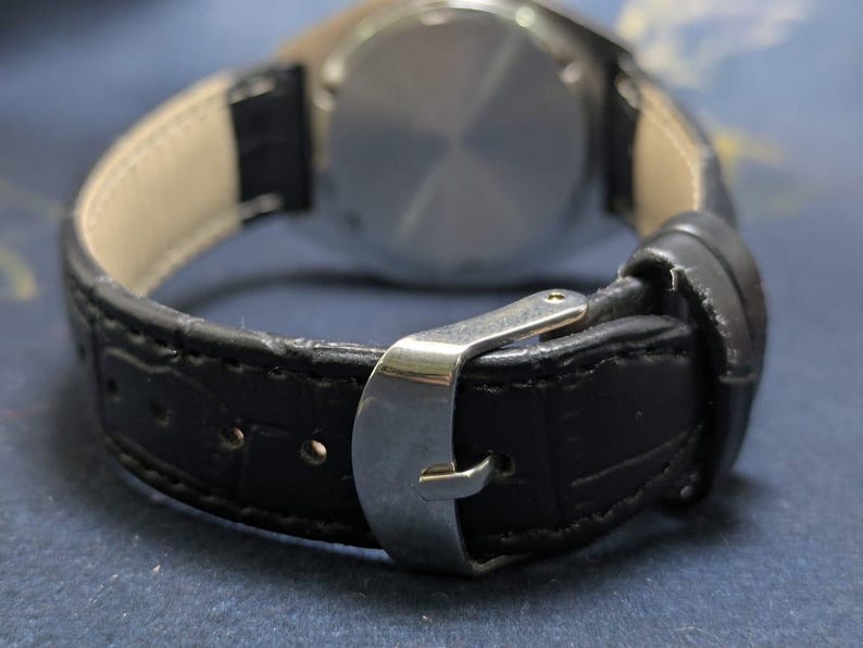 May include: Close-up of a black leather watch strap with a silver buckle. The watch strap is textured and stitched, with a light-colored interior. The watch face is visible in the background.