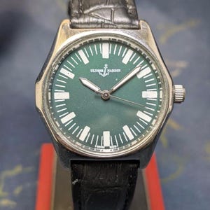 May include: A silver-toned wristwatch with a dark green face and a black leather band. The watch face features white hour markers and the brand name "ULYSSE NARDIN". The watch has a silver bezel and a crown on the side.