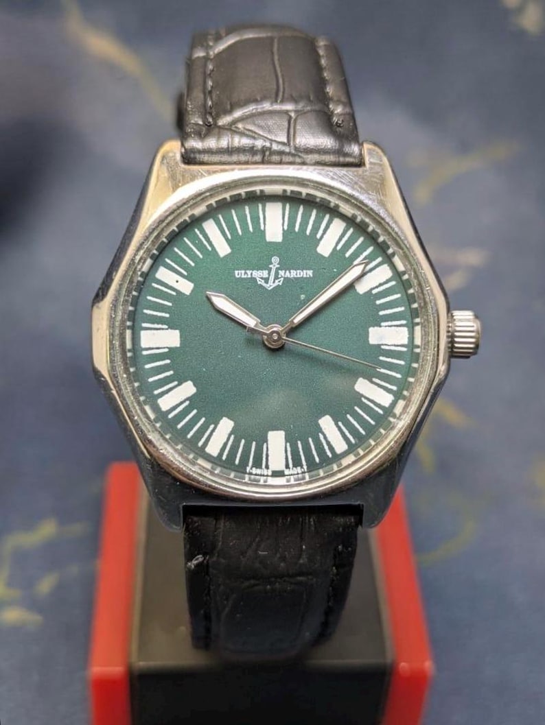 May include: A silver-toned wristwatch with a dark green face and a black leather band. The watch face features white hour markers and the brand name "ULYSSE NARDIN". The watch has a silver bezel and a crown on the side.