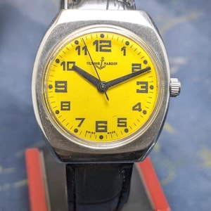 May include: A vintage Ulysse Nardin wristwatch with a bright yellow face and black hands. The watch has a silver-colored case and a black leather band. The watch face displays the numbers 1 through 12. The words "Ulysse Nardin" and "Swiss Made" are visible.