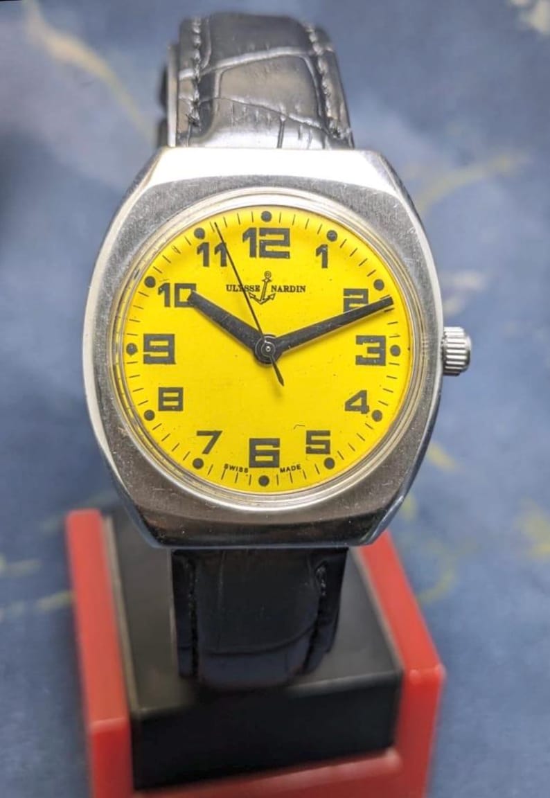 May include: A vintage Ulysse Nardin wristwatch with a bright yellow face and black hands. The watch has a silver-colored case and a black leather band. The watch face displays the numbers 1 through 12. The words "Ulysse Nardin" and "Swiss Made" are visible.